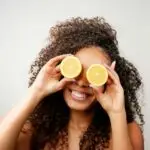 Smiling woman with curly hair playfully holds lemon slices in front of her eyes on a white background.